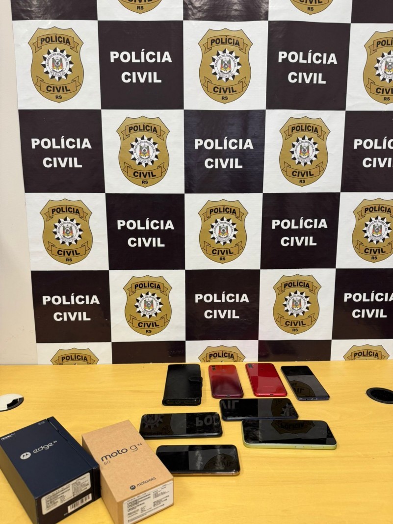A foto mostra vários celulares sob uma mesa e um banner da Polícia Civil ao fundo