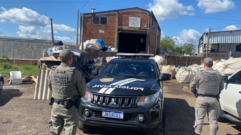 A foto mostra dois policiais militares e uma viatura da Polícia Civil em frente a um ponto de reciclagem