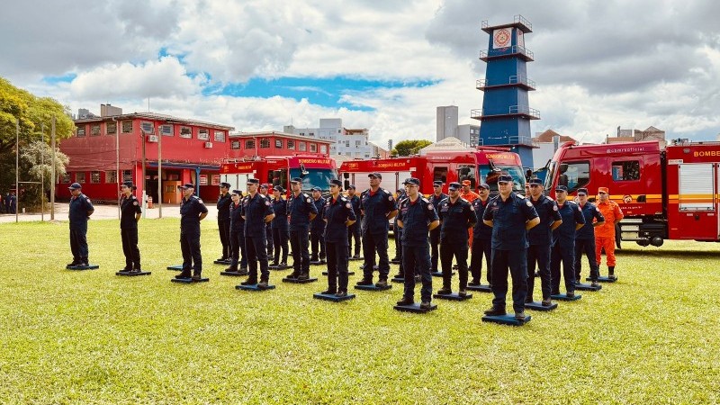 Imagem lateral mostra bombeiros militares de farda azul-escuro perfilados em frente a caminhões de bombeiro. Ao fundo, uma torre azul e dois prédios de cor vermelha, com um céu entre nuvens. 