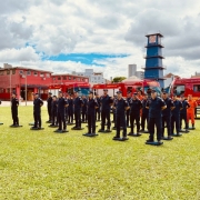 Imagem lateral mostra bombeiros militares de farda azul-escuro perfilados em frente a caminhões de bombeiro. Ao fundo, uma torre azul e dois prédios de cor vermelha, com um céu entre nuvens. 