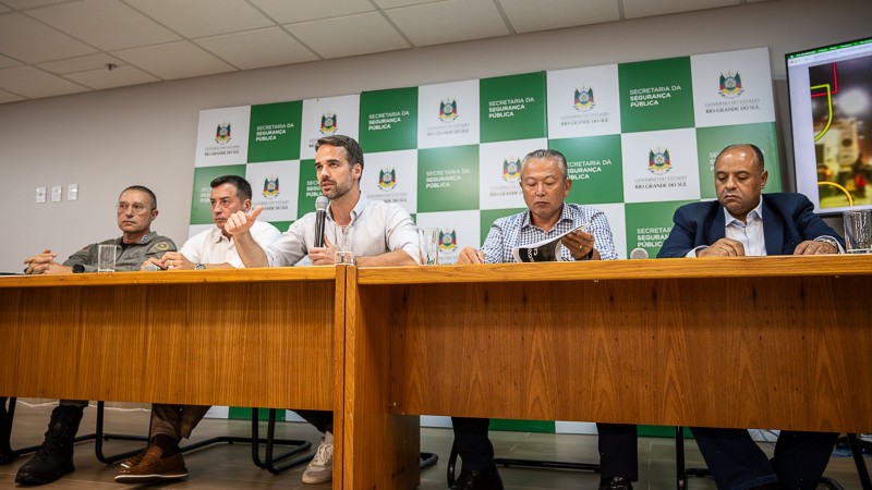 Autoridades sentados lado a lado em uma mesa, ao fundo o brasão do Estado