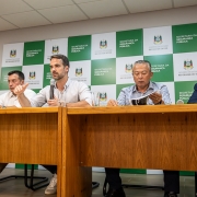 Autoridades sentados lado a lado em uma mesa, ao fundo o brasão do Estado