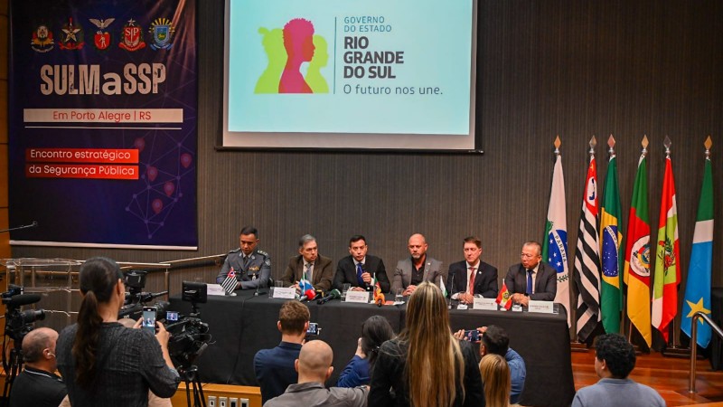 Os seis representantes das Secretarias de Segurança Pública estão sentados em uma mesa no palco do auditório. À frente, repórteres durante a coletiva de imprensa. Ao fundo, projetada, a logo do Governo do Estado do Rio Grande do Sul e o banner do evento, onde se lê "SULMaSSP Em Porto Alegre | RS" e "Encontro estratégico de Segurança Pública".