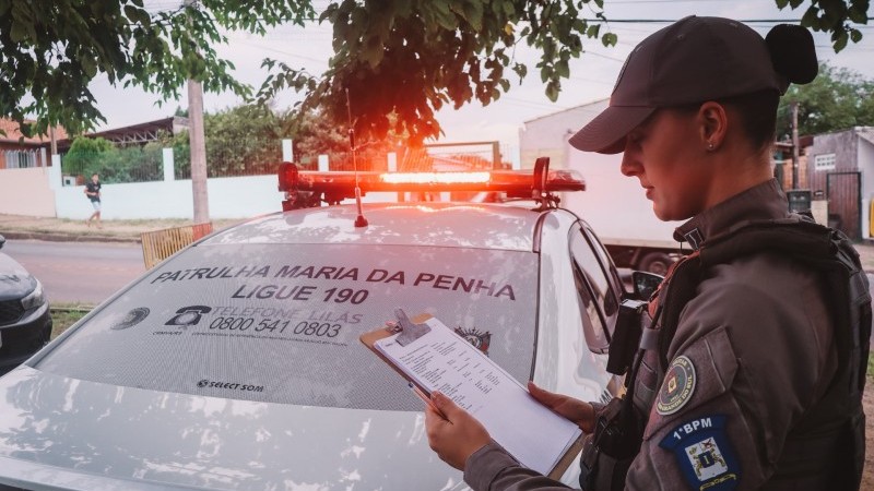 Imagem mostra uma policial da Patrulha Maria da Penha segurando uma planilha. Ao fundo há um carro da patrulha com as sirenes acessas