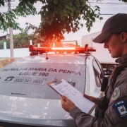 Imagem mostra uma policial  da Patrulha Maria da Penha segurando uma planilha. Ao fundo há um carro da patrulha com as sirenes acessas 
