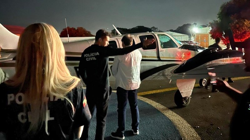 Policiais civis e o advogado suspeito sendo conduzido por hangar, em frente a uma aeronave