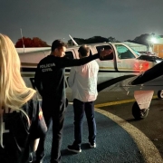 Policiais civis e o advogado suspeito sendo conduzido por hangar, em frente a uma aeronave