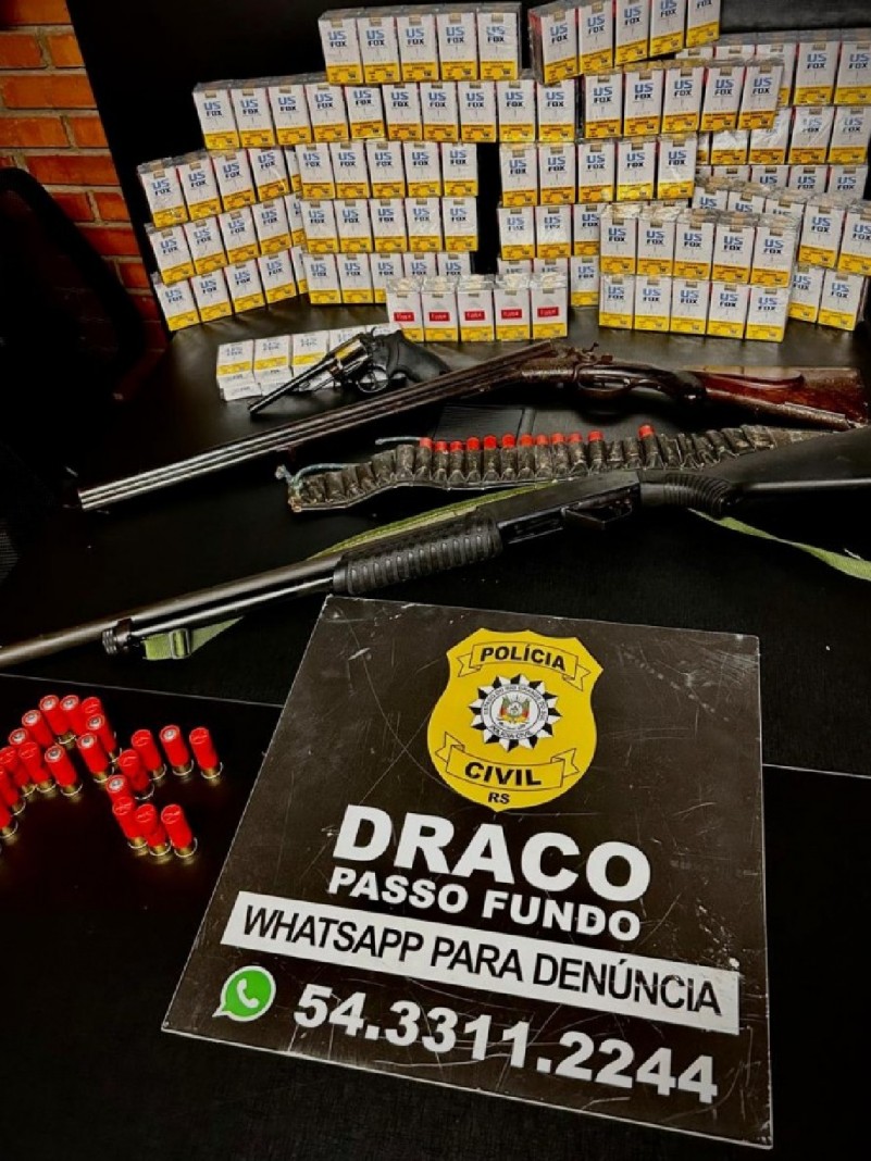 Cigarros, armas e munições apreendidos sobre uma mesa, com um cartaz do DRACO e um telefone WhatsApp para denúncias: 54 3311 2244