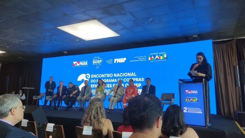 A foto, tirada da plateia, mostra o palco do local do evento. Há um telão com fundo azul sobre o qual se lê "2º Encontro Nacional do Programa de Compras Eficientes para o SUSP". À frente, há oito pessoas sentadas, voltadas para o público, e uma mulher fala no púlpito.