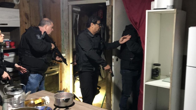 Policiais civis vasculham cômodo em uma casa. Panelas e outros utensílios de cozinha estão sobre uma mesa. Agentes fardados levantam uma cortina para acessar o cômodo anexo.