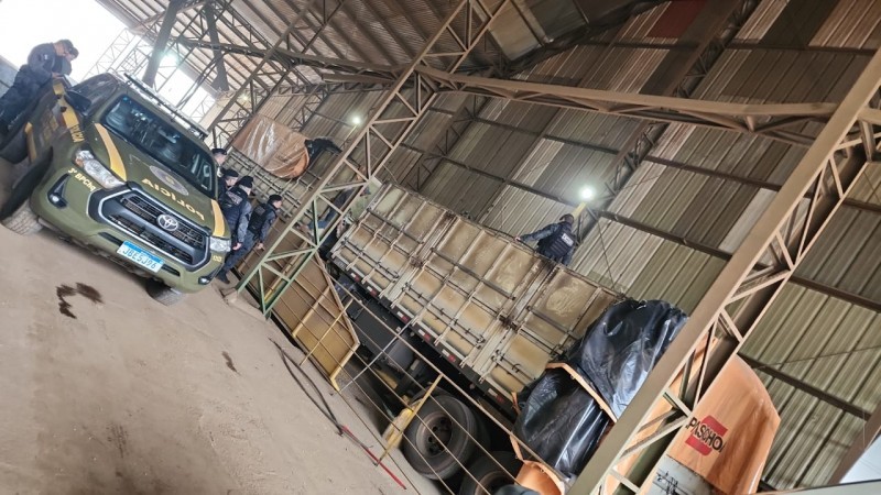 A imagem mostra um caminhão apreendido dentro de um depósito. Também estão na imagem agentes da Brigada Militar e uma viatura da corporação