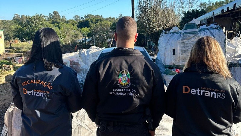 A imagem mostra três servidores das forças da Segurança Pública do Estado em um dos locais fiscalizados pela operação. No lado direito está uma mulher com jaqueta do DetranRS. No centro, há um homem com uma jaqueta da SSP. Na esquerda, há uma mulher com jaqueta do Detran/RS. Os três estão de costas para a câmera.
