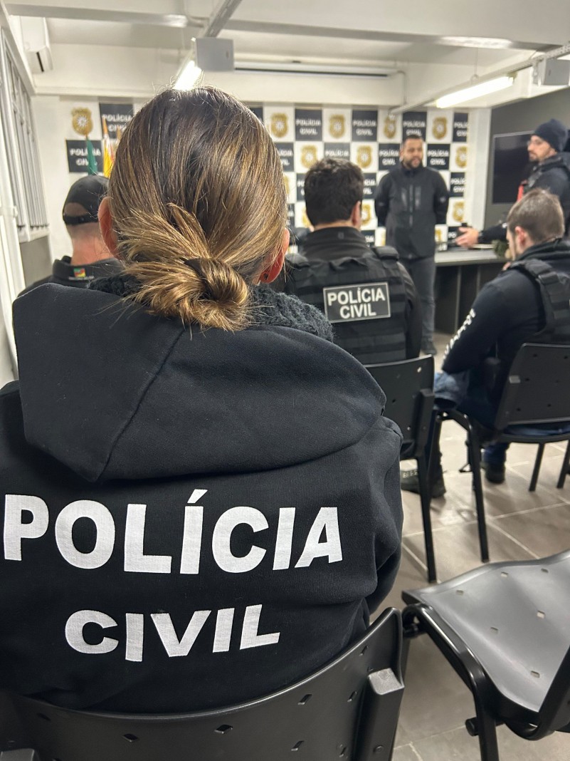 A foto mostra agentes da Polícia Civil de costas, com o nome da corporação em seus uniformes. Eles estão sentados em uma sala, disposta no formato auditório, e à frente, dois policiais falam voltados para os demais. Há um fundo de palco quadriculado com imagens alternadas do logotipo da Polícia Civil e o nome da instituição.
