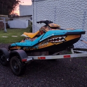 Uma moto aquática nas cores laranja e azul, com um adesivo grande de dentes pontiagudos fixado na lateral inferior, está em um reboque, nos fundos de uma residência.
