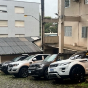 Quatro automóveis tipo SUV, dois brancos, um preto e um chumbo, estão estacionados lado a lado em um terreno íngreme.
