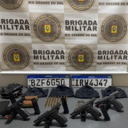 A imagem mostra pistolas, munições, balaclavas e placas de automóveis em uma mesa. Ao fundo, há um banner da Brigada Militar