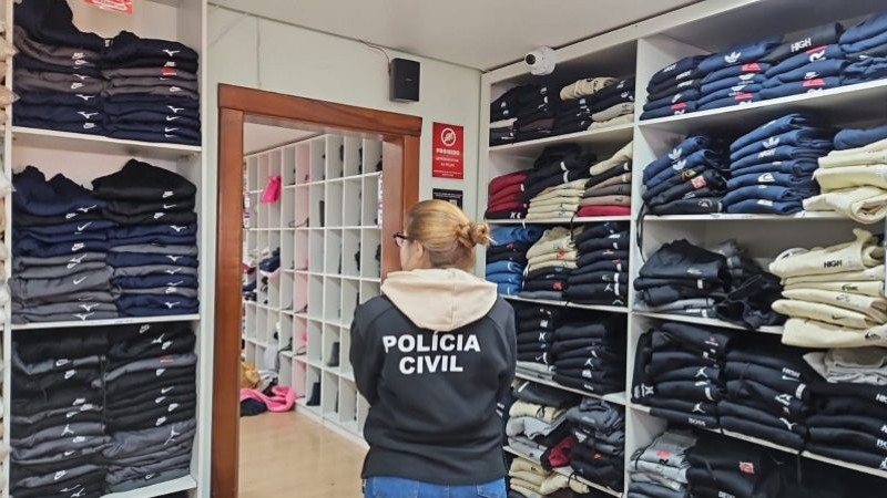 A imagem mostra uma agente da polícia civil em meio aos materiais apreendidos. A agente usa uma jaqueta da polícia civil e está de costas para a câmera. Ao redor dela, estão roupas empilhadas em estantes. À frente dela há uma porta aberta que dá para uma outra sala, onde estão outros materiais apreendidos