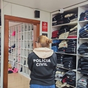 A imagem mostra uma agente da polícia civil em meio aos materiais apreendidos. A agente usa uma jaqueta da polícia civil e está de costas para a câmera. Ao redor dela, estão roupas empilhadas em estantes. À frente dela há uma porta aberta que dá para uma outra sala, onde estão outros materiais apreendidos