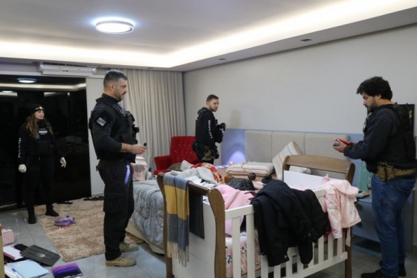 Quatro agentes da Polícia Civil cumprem mandados judiciais em um quarto. No primeiro plano, há um berço com roupas e cobertas atiradas.
