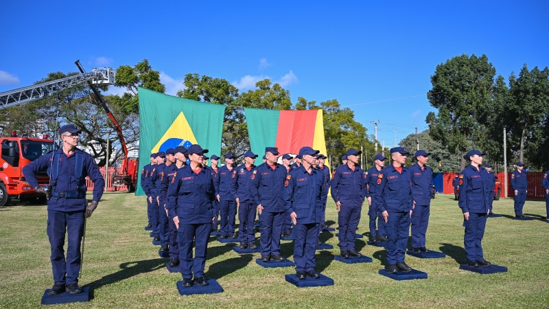 A imagem retrata uma cerimônia de formatura do Corpo de Bombeiros Militar do Rio Grande do Sul. Bombeiros uniformizados estão perfilados em um gramado, com bandeiras do Brasil e do RS ao fundo, além de um caminhão e equipamentos de resgate visíveis. O cenário é ao ar livre, com céu limpo e árvores ao redor, transmitindo um clima solene e de celebração da conquista profissional.