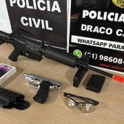 Sobre uma mesa de madeira, objetos apreendidos: aparelhos celulares, cartões bancários e simulacros de pistola e fuzil. Ao fundo, cartazes da Polícia Civil com o escudo, um telefone whatsapp que serve para denúncia: 51 98608 9984