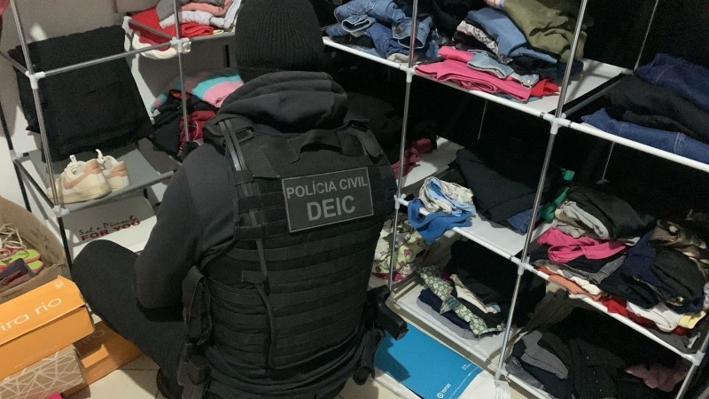 Agente fardado com identificação do DEIC vasculha prateleiras de roupas e calçados