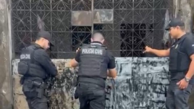 Três agentes da Polícia Civil cumprem mandados: três homens forçam a entrada em um edifício, pressionando grade de metal acinzentado 