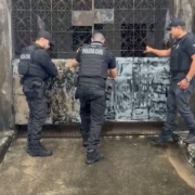 Três agentes da Polícia Civil cumprem mandados: três homens forçam a entrada em um edifício, pressionando grade de metal acinzentado 