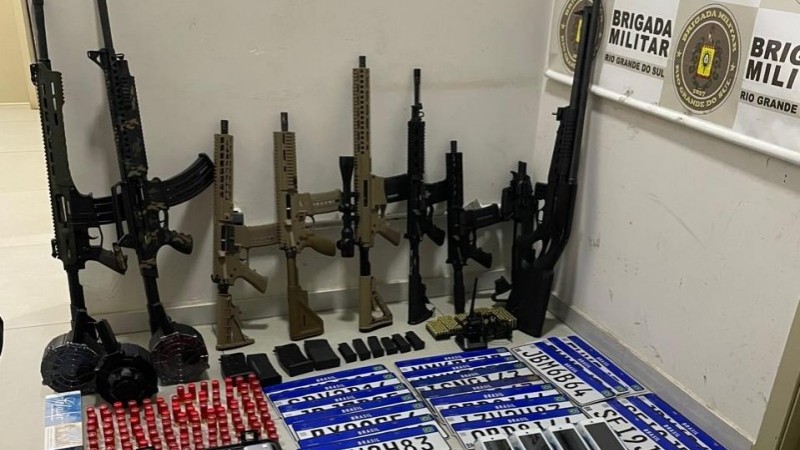 A imagem mostra armas de grosso calibre, como fuzis, carabinas, escopetas e espingardas apreendidas pela BM. Elas estão posicionadas ao fundo da imagem, encostadas em uma parede. Também encontram-se na imagem munições, placas de veículos, celulares, balanças de precisão, máquinas de cartão, papelotes de maconha, cocaína e crack, além de notas de 50, 20, dez, cinco e dois reais