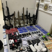 A imagem mostra armas de grosso calibre, como fuzis, carabinas, escopetas e espingardas apreendidas pela BM. Elas estão posicionadas ao fundo da imagem, encostadas em uma parede. Também encontram-se na imagem munições, placas de veículos, celulares, balanças de precisão, máquinas de cartão, papelotes de maconha, cocaína e crack, além de notas de 50, 20, dez, cinco e dois reais
