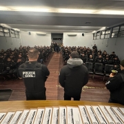 A imagem mostra três agentes da Polícia Civil de costas para a câmera e de frente para uma plateia composta por outros agentes. Os três policiais estão de pé, enquanto os demais agentes estão sentados. Ao fundo da sala, há algumas pessoas em pé