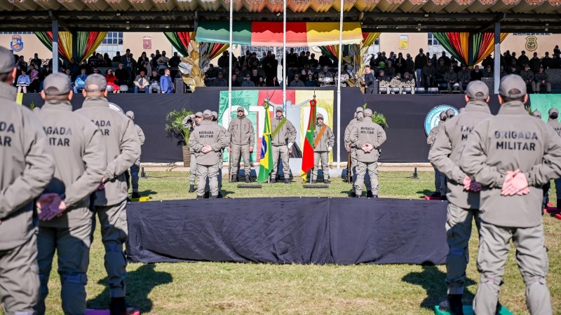 A imagem mostra os formandos de frente para a tribuna principal, em posição de descanso, durante a solenidade de formatura. Eles usam fardas da Brigada Militar e boné