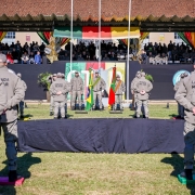 A imagem mostra os formandos de frente para a tribuna principal, em posição de descanso, durante a solenidade de formatura. Eles usam fardas da Brigada Militar e boné 