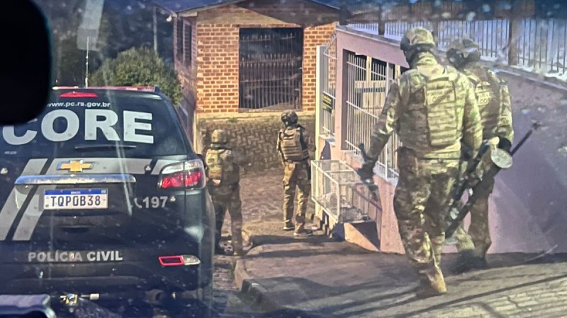 Imagem mostra quatro policiais armados descendo uma rua para realizar uma abordagem. Ao lado, uma viatura estacionada