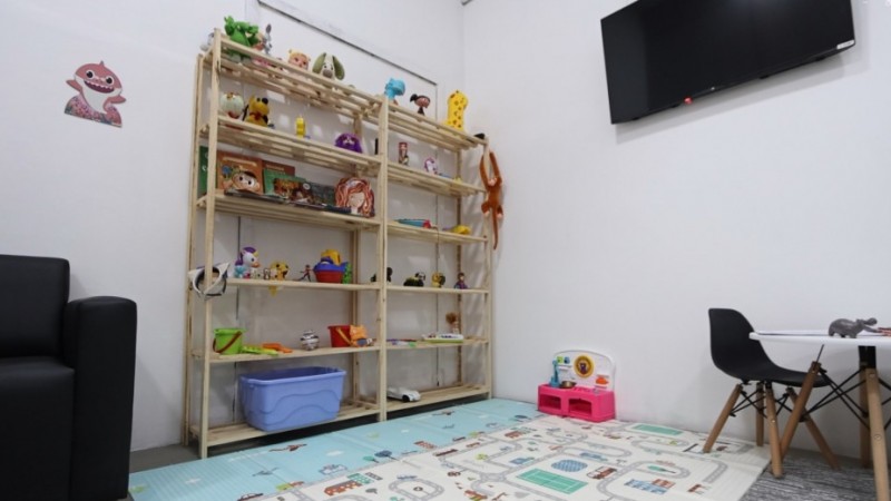 Sala infantil com ambiente acolhedor e brinquedos. &Agrave; esquerda, h&aacute; parte de um sof&aacute; preto. No centro, uma estante de madeira exibe diversos brinquedos e livros infantis. No ch&atilde;o, um tapete colorido com desenhos de ruas e pistas cobre parte do espa&ccedil;o. &Agrave; direita, h&aacute; uma pequena mesa branca com duas cadeiras infantis e brinquedos sobre o tampo. Na parede, acima da estante, h&aacute; uma televis&atilde;o preta desligada. O ambiente &eacute; bem iluminado, com paredes brancas e apar&ecirc;ncia organizada e l&uacute;dica.