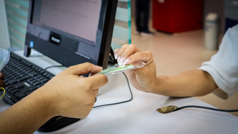 Foto de uma banca de atendimento, com um computador ao fundo. Em destaque est&aacute; uma m&atilde;o entregando uma carteira de identidade a outra, que pega o documento.