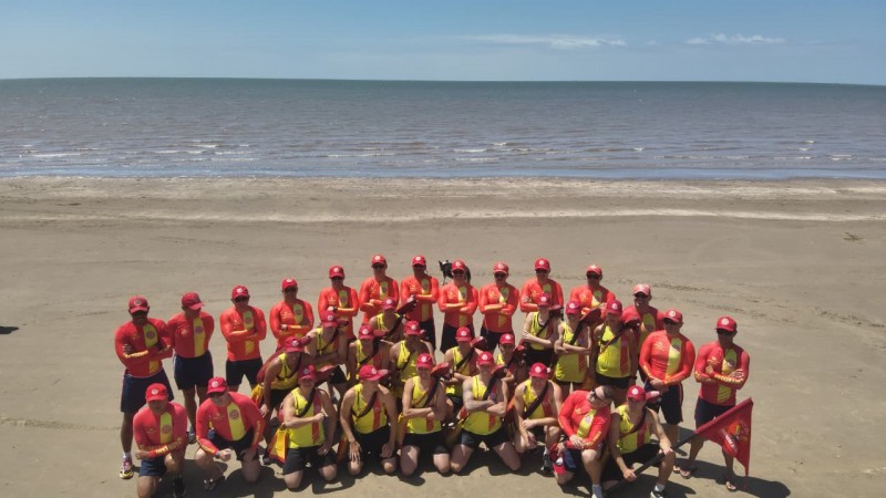 A foto mostra um grande grupo de guarda-vidas reunidos para uma foto coletiva na praia. Todos vestem uniformes vermelhos e amarelos, com bon&eacute;s vermelhos. Ao fundo, h&aacute; o mar e um horizonte limpo sob c&eacute;u azul claro.