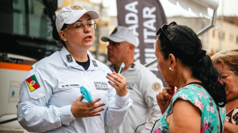 A imagem mostra uma policial do Departamento de Sa&uacute;de da Brigada Militar conversando com uma mulher. Ambas est&atilde;o pr&oacute;ximas a uma base m&oacute;vel do projeto Pol&iacute;cia de Proximidade. &Eacute; poss&iacute;vel ver ao fundo outro policial do Departamento de Sa&uacute;de da Brigada Militar, al&eacute;m de outra mulher