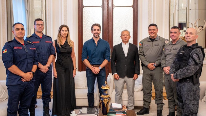 Esta &eacute; uma descri&ccedil;&atilde;o detalhada da imagem para fins de acessibilidade: Vis&atilde;o Geral A imagem registra um momento oficial em que oito pessoas &mdash; sete homens e uma mulher &mdash; posam para uma fotografia em um ambiente luxuoso e formal. O grupo est&aacute; organizado em uma linha reta, transmitindo uma atmosfera de coopera&ccedil;&atilde;o institucional. Pessoas e Vestimentas (da esquerda para a direita) Bombeiros Militares: Os dois primeiros homens &agrave; esquerda vestem uniformes operacionais azul-marinho com detalhes e identifica&ccedil;&otilde;es em vermelho, t&iacute;picos do Corpo de Bombeiros. Civil: Uma mulher com cabelos loiros longos veste um vestido preto longo e elegante. Governador: No centro da imagem, Eduardo Leite veste uma camisa social azul-marinho com as mangas levemente dobradas e cal&ccedil;a jeans escura. Civil/Autoridade: &Agrave; direita do governador, um homem de cabelos grisalhos veste um blazer preto, camisa branca e cal&ccedil;a cinza-clara. Brigada Militar: Os dois homens seguintes vestem fardas cinza-oliva da Brigada Militar, com diversos distintivos e ins&iacute;gnias. T&aacute;tico: Na extremidade direita, um homem veste um uniforme t&aacute;tico camuflado em tons de cinza e preto, equipado com colete operacional. Cen&aacute;rio e Composi&ccedil;&atilde;o O Ambiente: O encontro ocorre em uma sala de recep&ccedil;&atilde;o cl&aacute;ssica, possivelmente no Pal&aacute;cio Piratini. O fundo apresenta uma grande porta dupla de madeira com pain&eacute;is de vidro fosco, paredes brancas decoradas com boiseries (molduras de gesso) e espelhos laterais. Mobili&aacute;rio: Nas laterais, veem-se partes de sof&aacute;s brancos. &Agrave; frente do grupo, h&aacute; uma mesa de centro de madeira escura contendo livros, uma pequena x&iacute;cara de caf&eacute; sobre um pires branco e um vaso decorativo alongado em azul-marinho com detalhes dourados. Ilumina&ccedil;&atilde;o: A luz &eacute; suave e bem distribu&iacute;da, real&ccedil;ando os detalhes dos uniformes e do ambiente.
