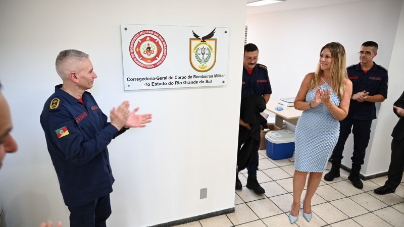 Em frente a uma parede branca, uma placa institucional j&aacute; descoberta est&aacute; fixada, com os bras&otilde;es e o texto da Corregedoria-Geral do Corpo de Bombeiros Militar do Estado do Rio Grande do Sul. Um bombeiro militar est&aacute; &agrave; esquerda, aplaudindo. &Agrave; direita, uma mulher de vestido claro e sapatos de salto tamb&eacute;m aplaude. Atr&aacute;s dela, outros bombeiros acompanham a cena. O ambiente &eacute; interno, organizado e bem iluminado, transmitindo um clima formal e comemorativo.