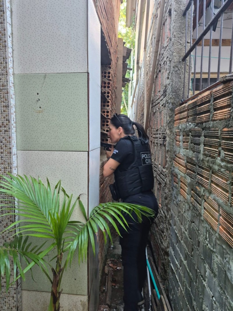 Uma pessoa vestida com uniforme escuro e colete t&aacute;tico da Pol&iacute;cia Civil est&aacute; posicionada em um espa&ccedil;o muito estreito entre duas constru&ccedil;&otilde;es. O corredor &eacute; apertado, com paredes de alvenaria dos dois lados, feitas de tijolos aparentes e concreto, mostrando textura irregular e sinais de desgaste.
A pessoa est&aacute; de perfil, com o corpo levemente inclinado para frente, apoiando-se pr&oacute;xima &agrave; parede da esquerda, como se observasse ou verificasse algo &agrave; frente. O uniforme possui identifica&ccedil;&atilde;o escrita nas costas. Os bra&ccedil;os est&atilde;o pr&oacute;ximos ao corpo, e o espa&ccedil;o limita bastante os movimentos.
No primeiro plano, &agrave; esquerda, h&aacute; uma planta ornamental com folhas longas e verdes, contrastando com o ambiente urbano e fechado. No ch&atilde;o do corredor, v&ecirc;-se terra, pequenos detritos e alguns canos ou mangueiras encostados na parede. Ao fundo, entra um pouco de luz natural, sugerindo uma abertura maior adiante.