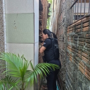 Uma pessoa vestida com uniforme escuro e colete t&aacute;tico da Pol&iacute;cia Civil est&aacute; posicionada em um espa&ccedil;o muito estreito entre duas constru&ccedil;&otilde;es. O corredor &eacute; apertado, com paredes de alvenaria dos dois lados, feitas de tijolos aparentes e concreto, mostrando textura irregular e sinais de desgaste.
A pessoa est&aacute; de perfil, com o corpo levemente inclinado para frente, apoiando-se pr&oacute;xima &agrave; parede da esquerda, como se observasse ou verificasse algo &agrave; frente. O uniforme possui identifica&ccedil;&atilde;o escrita nas costas. Os bra&ccedil;os est&atilde;o pr&oacute;ximos ao corpo, e o espa&ccedil;o limita bastante os movimentos.
No primeiro plano, &agrave; esquerda, h&aacute; uma planta ornamental com folhas longas e verdes, contrastando com o ambiente urbano e fechado. No ch&atilde;o do corredor, v&ecirc;-se terra, pequenos detritos e alguns canos ou mangueiras encostados na parede. Ao fundo, entra um pouco de luz natural, sugerindo uma abertura maior adiante.