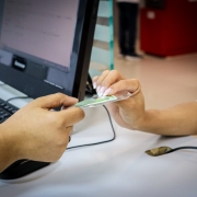 Em primeiro plano, uma pessoa est&aacute; entregando um cart&atilde;o plastificado, um documento de identidade, para outra pessoa. O cart&atilde;o &eacute; segurado com a m&atilde;o estendida, sugerindo um momento de verifica&ccedil;&atilde;o ou registro. Ao fundo, h&aacute; um computador de mesa com teclado e monitor, refor&ccedil;ando o contexto administrativo ou oficial da intera&ccedil;&atilde;o. O cen&aacute;rio transmite a ideia de um processo formal de atendimento ou confer&ecirc;ncia de documentos.