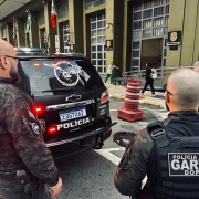 Tr&ecirc;s policiais da unidade GARRA, da Pol&iacute;cia Civil, aparecem em frente a uma viatura preta com emblemas da corpora&ccedil;&atilde;o e da unidade especial. Eles vestem uniformes camuflados e equipamentos t&aacute;ticos. Ao fundo, h&aacute; um pr&eacute;dio oficial da Pol&iacute;cia Civil, bandeiras do Brasil e de S&atilde;o Paulo, al&eacute;m de pessoas circulando, indicando um ambiente urbano.