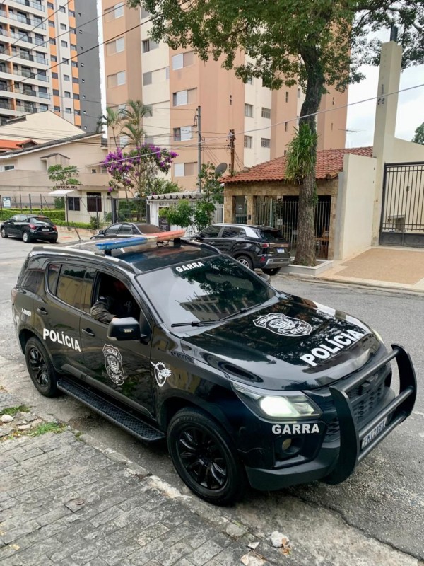 Um SUV preto da Polícia Civil está estacionado em uma rua de bairro. O veículo tem luzes de emergência no teto, rodas pretas e para-choque reforçado. As inscrições “POLÍCIA” e “GARRA” aparecem em destaque na lataria. O entorno mostra prédios residenciais, árvores e outros carros estacionados, caracterizando uma área urbana comum.