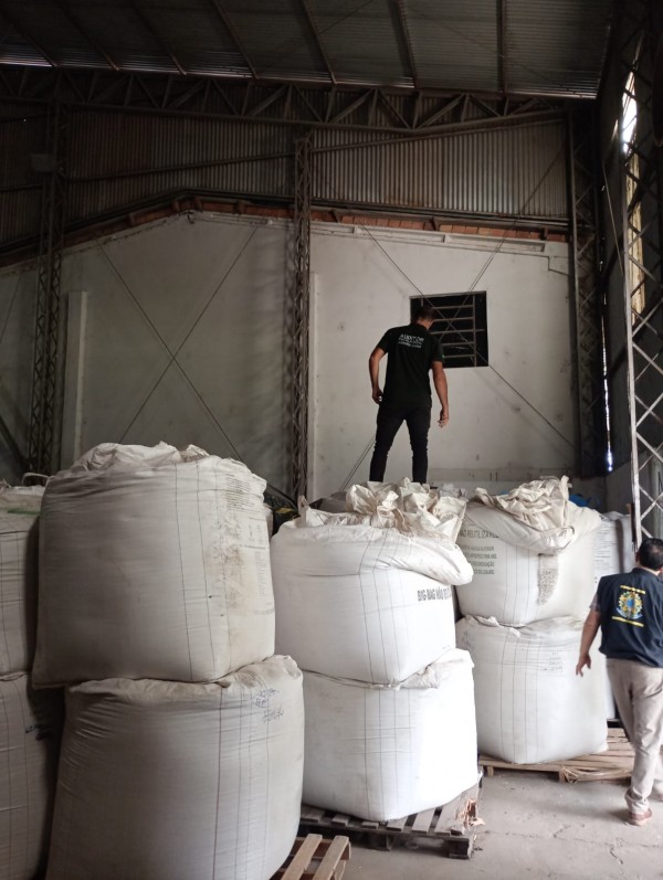 Sacos de sementes que apresentaram irregularidades. Um galpão amplo, com teto de metal. No centro, grandes sacos brancos empilhados sobre paletes de madeira. Um homem está em pé sobre os sacos, usando camiseta branca com a frase “Soluções Ambientais” nas costas. Outro homem caminha no chão, ao lado das pilhas. Ao fundo, há uma pequena janela na parede.