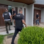 Exterior de uma casa moderna. H&aacute; um caminho de piso claro e um arbusto verde ao lado da entrada. V&aacute;rios policiais, com coletes pretos escritos &ldquo;POL&Iacute;CIA&rdquo;, aproximam-se da porta. Um deles usa uma ferramenta para abrir a porta, enquanto outros observam e se preparam para entrar. A cena mostra uma opera&ccedil;&atilde;o policial em andamento. 