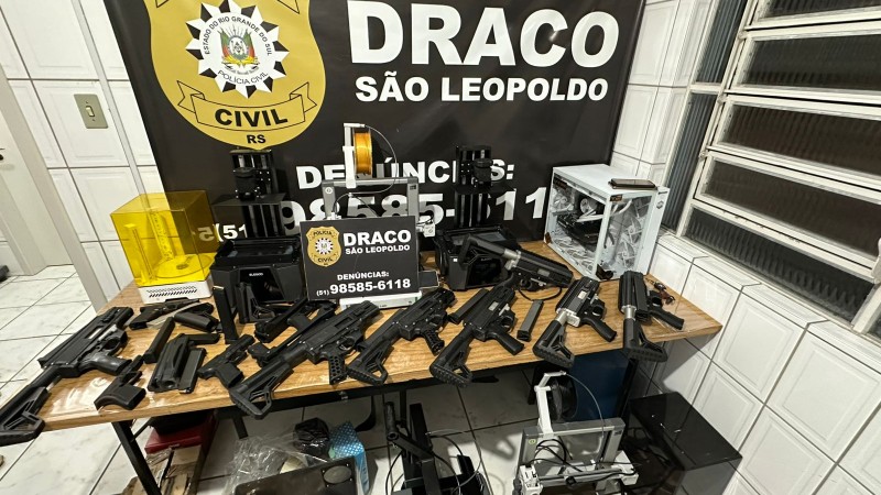 A imagem mostra uma opera&ccedil;&atilde;o da Pol&iacute;cia Civil do Rio Grande do Sul, realizada pela unidade DRACO de S&atilde;o Leopoldo. Em destaque, h&aacute; um banner institucional com o nome da corpora&ccedil;&atilde;o e um n&uacute;mero de contato para den&uacute;ncias. &Agrave; frente do banner, sobre uma mesa, est&atilde;o expostos diversos itens apreendidos: armas de fogo, componentes de armas e v&aacute;rias impressoras 3D, sugerindo que a tecnologia estava sendo utilizada para fabricar ou modificar armamentos. O cen&aacute;rio transmite claramente o car&aacute;ter de uma a&ccedil;&atilde;o policial voltada ao combate &agrave; produ&ccedil;&atilde;o e distribui&ccedil;&atilde;o ilegal de armas.