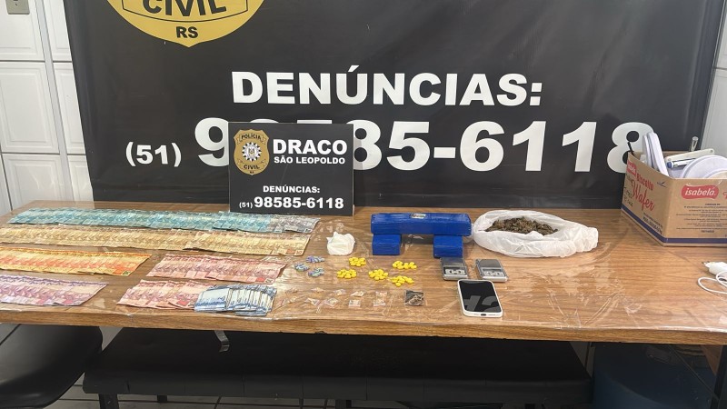 A imagem retrata uma apreens&atilde;o policial realizada pela Pol&iacute;cia Civil do Rio Grande do Sul, onde sobre uma mesa est&atilde;o expostos dinheiro em esp&eacute;cie, drogas embaladas em diferentes formatos, comprimidos, balan&ccedil;as digitais e um celular, diante de um banner da DRACO S&atilde;o Leopoldo com informa&ccedil;&otilde;es de contato para den&uacute;ncias: 51 98585 6118.