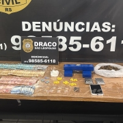 A imagem retrata uma apreens&atilde;o policial realizada pela Pol&iacute;cia Civil do Rio Grande do Sul, onde sobre uma mesa est&atilde;o expostos dinheiro em esp&eacute;cie, drogas embaladas em diferentes formatos, comprimidos, balan&ccedil;as digitais e um celular, diante de um banner da DRACO S&atilde;o Leopoldo com informa&ccedil;&otilde;es de contato para den&uacute;ncias: 51 98585 6118.